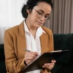 Businesswoman in a brown suit writing on clipboard indoors, captured in a professional setting.